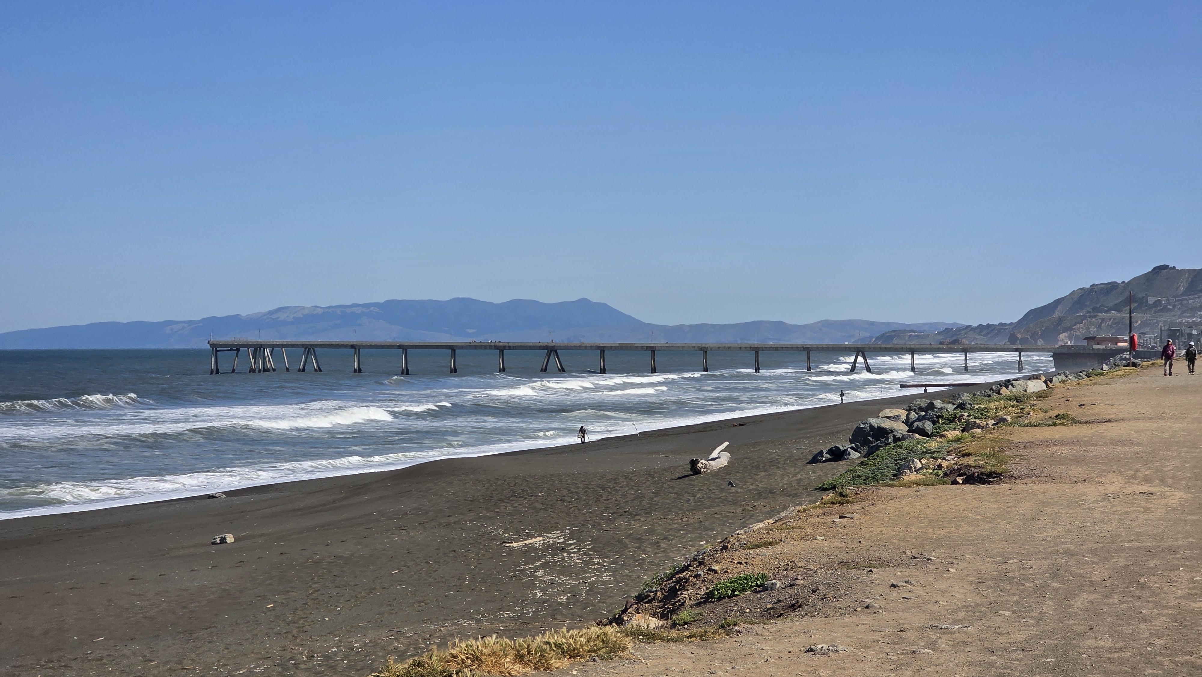 Walkers on the path beside the Pacifica Pier and black sand beach